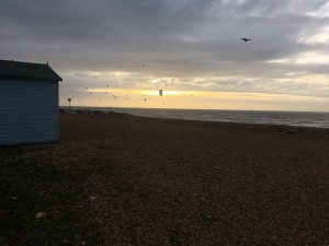 Pebbles, sea with many kitesurfers, one blue beach hut and cloudy sky with orange sunlight breaking through