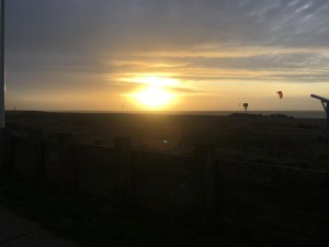 Sunrise over beach, with a kitesurfer in the distance and the wooden fencing at the front, pebbles in between