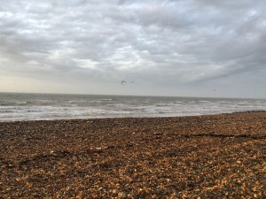 Beach with pebbles and rough grey sea, clouds above, a kitesurfer in the sea