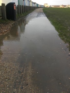 Huge puddle spreading across path, with grass to the right and beach huts to the left