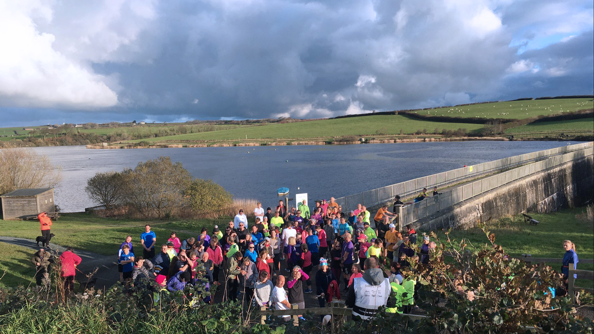 A Run Director in the foreground gives a briefinng to runners assembled in front of him in brightly coloured clothes with the expanse of Tamar Lake in the behind them all.